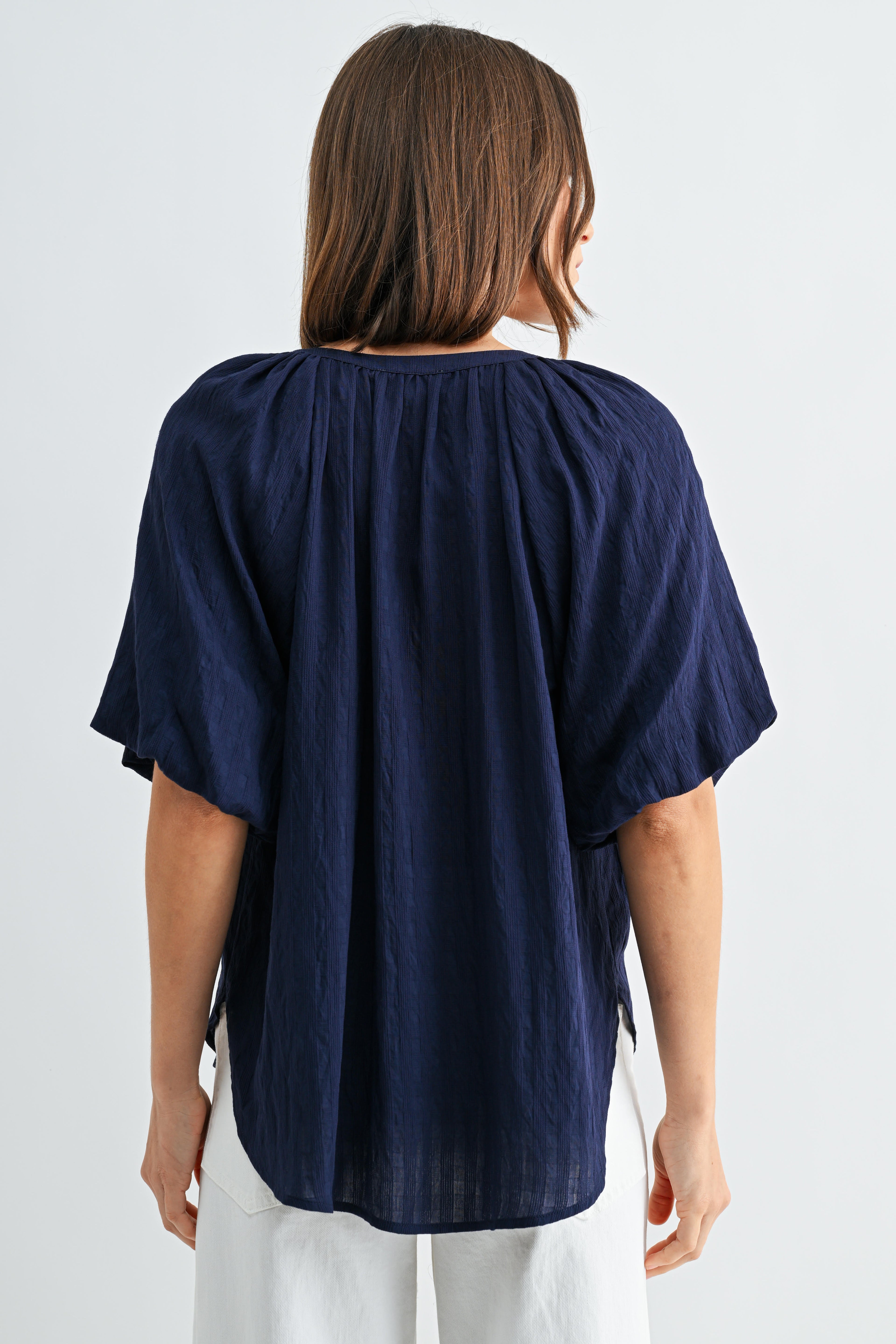 Back view of a woman wearing a textured navy blue blouse with short balloon sleeves with white buttons on a light gray background