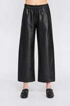Black wide barrel leg faux leather pants with an elastic waistband, shown on a model wearing black loafers.