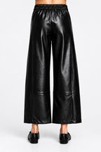Back view of a black wide barrel leg faux leather pants with an elastic waistband, shown on a model wearing black loafers.