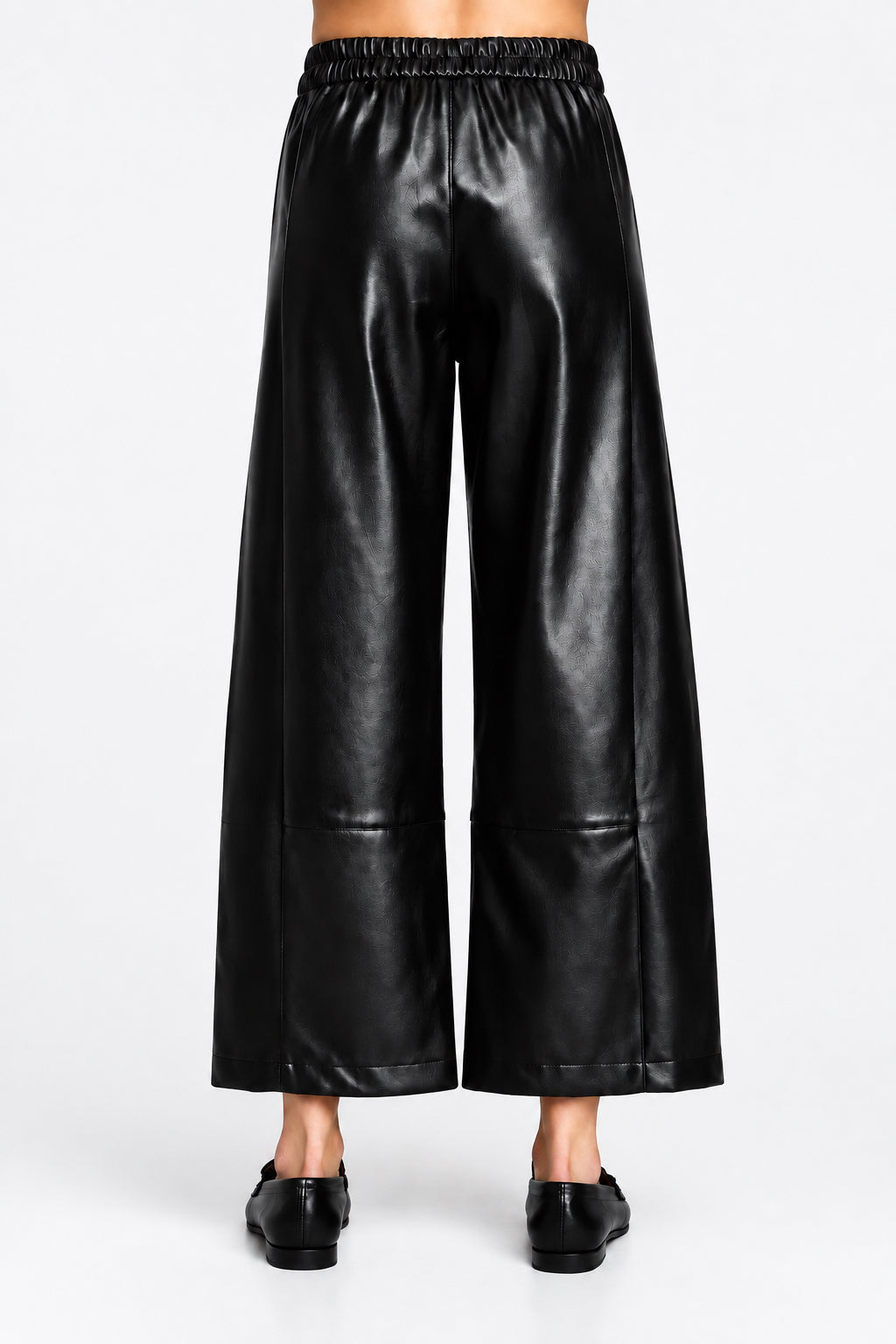 Back view of a black wide barrel leg faux leather pants with an elastic waistband, shown on a model wearing black loafers.