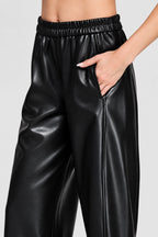Black wide barrel leg faux leather pants with an elastic waistband and side pockets shown on a model wearing black loafers.
