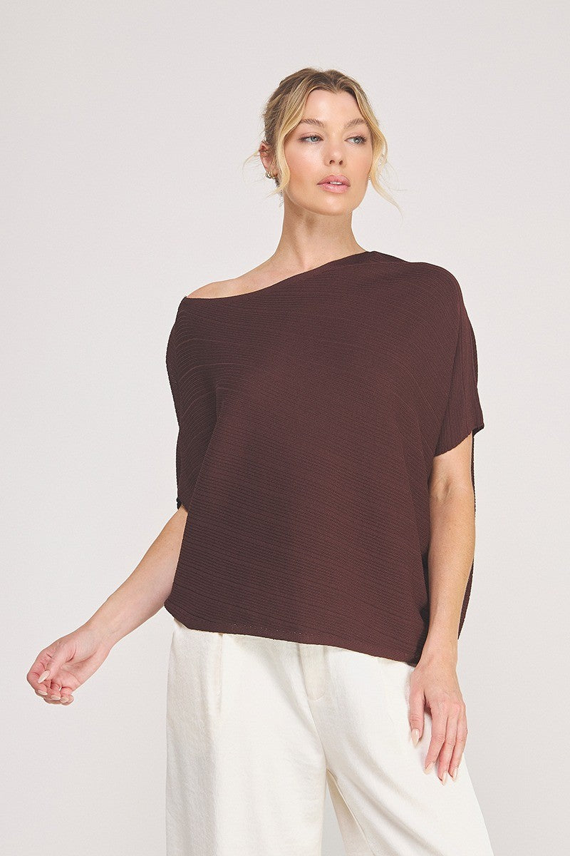 A woman wearing an off the shoulder boat neck top in espresso with ribbed knit and asymmetrical hemline against a white background with white pants