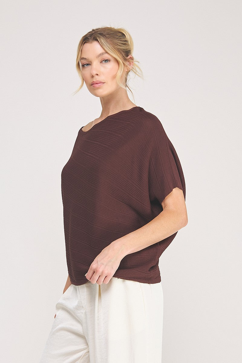 A woman wearing an off the shoulder boat neck top in espresso with ribbed knit and asymmetrical hemline against a white background with white pants