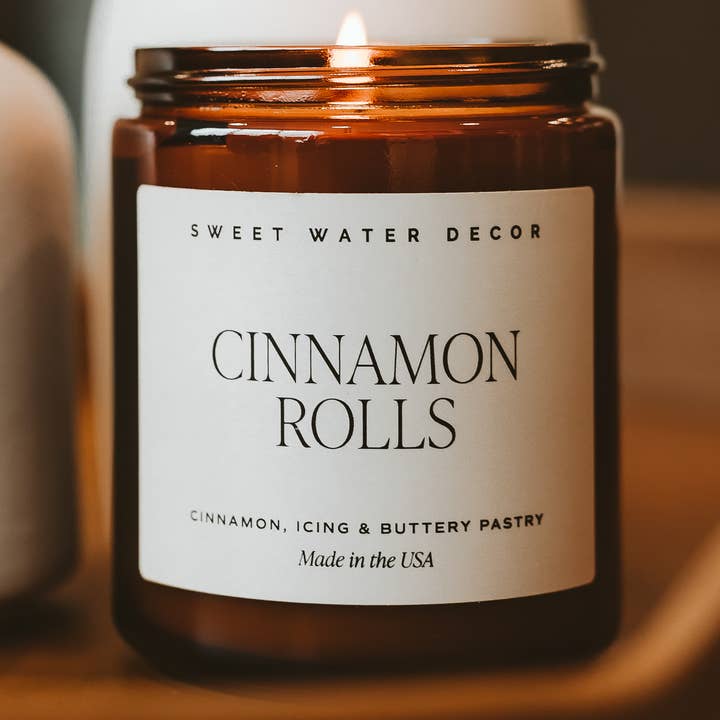 An amber glass jar candle with a white label reading 'SWEET WATER DECOR' and 'CINNAMON ROLLS', photographed close-up on a warm wooden surface.