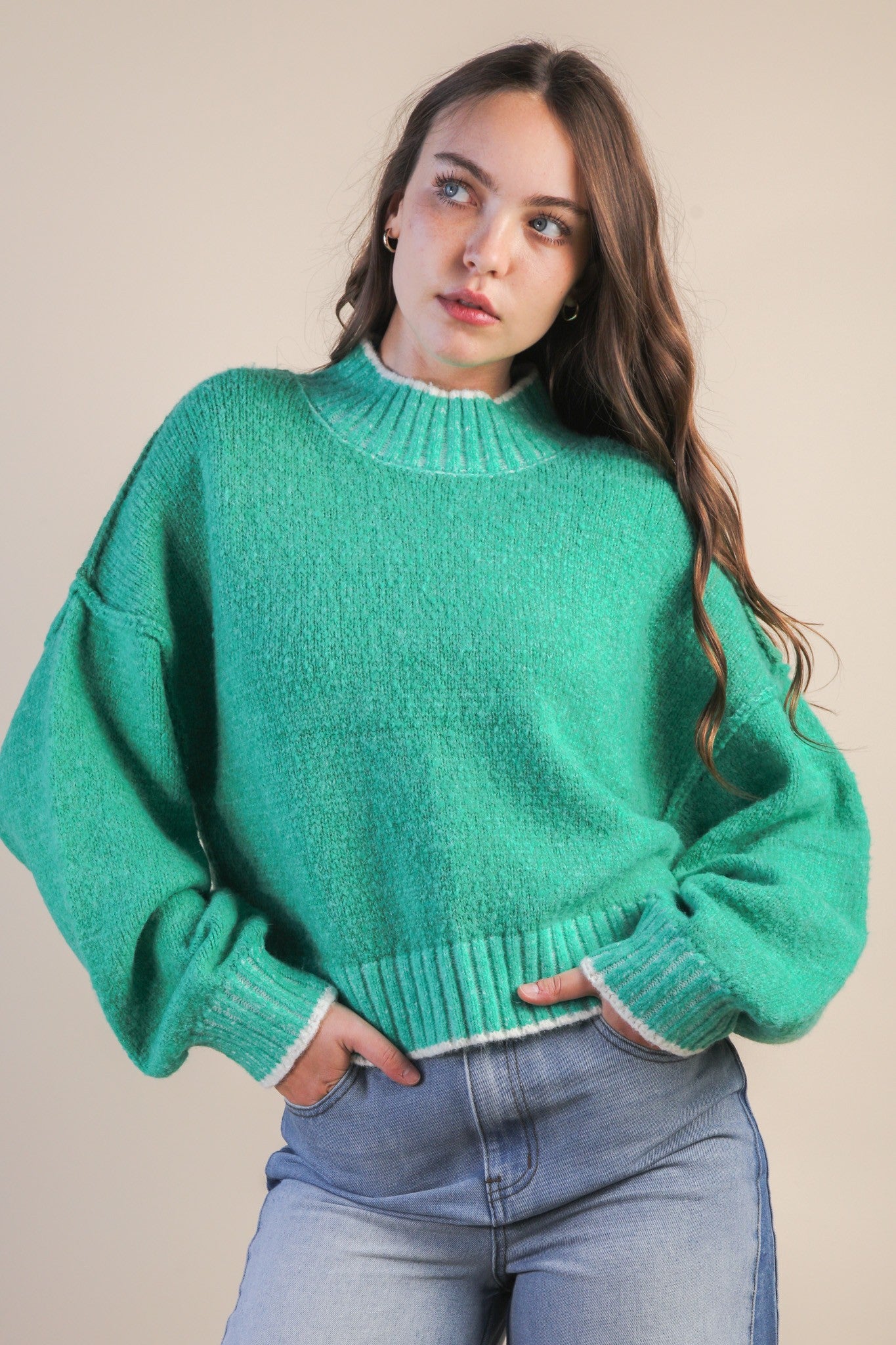 Woman wearing a green mock neck knit sweater with white trim and blue jeans against a beige background