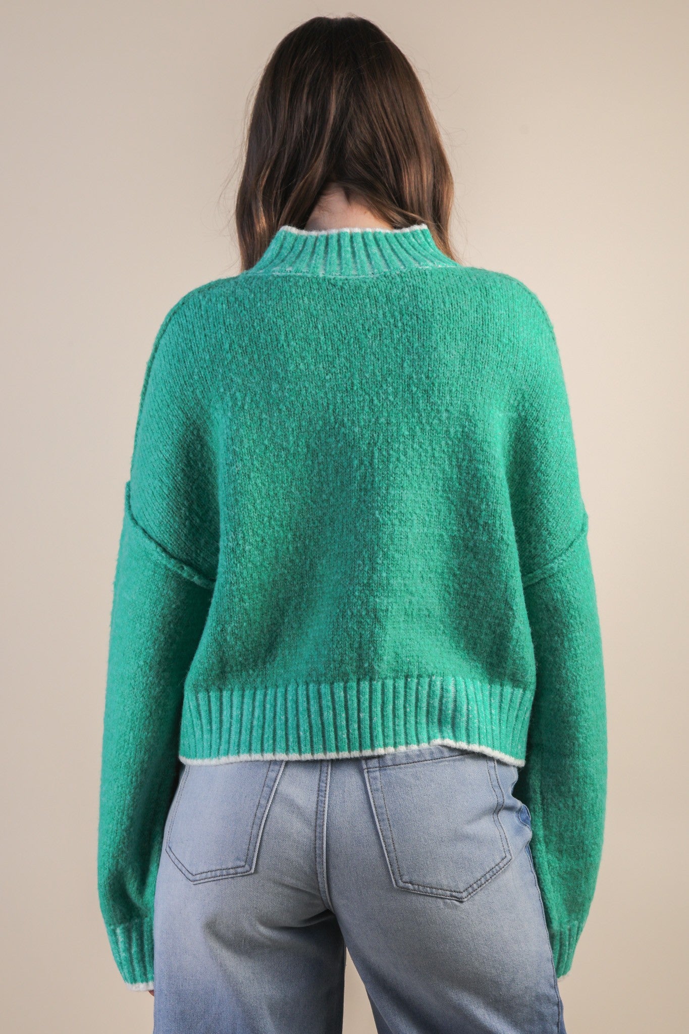 Back view of woman wearing a green mock neck knit sweater with white trim and blue jeans against a beige background