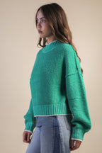 Woman wearing a green mock neck knit sweater with white trim and blue jeans against a beige background