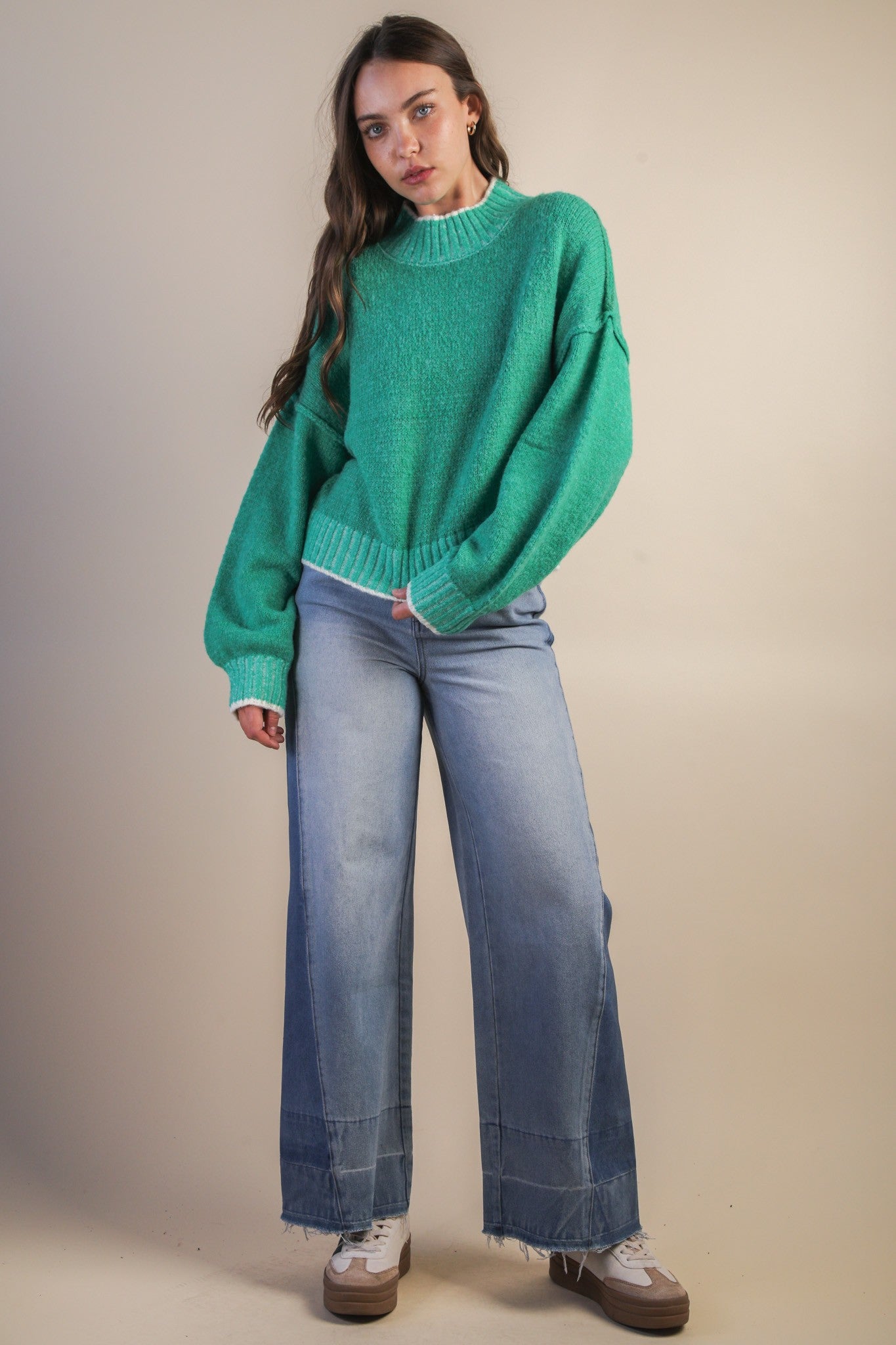 Woman wearing a green mock neck knit sweater with white trim and blue jeans against a beige background