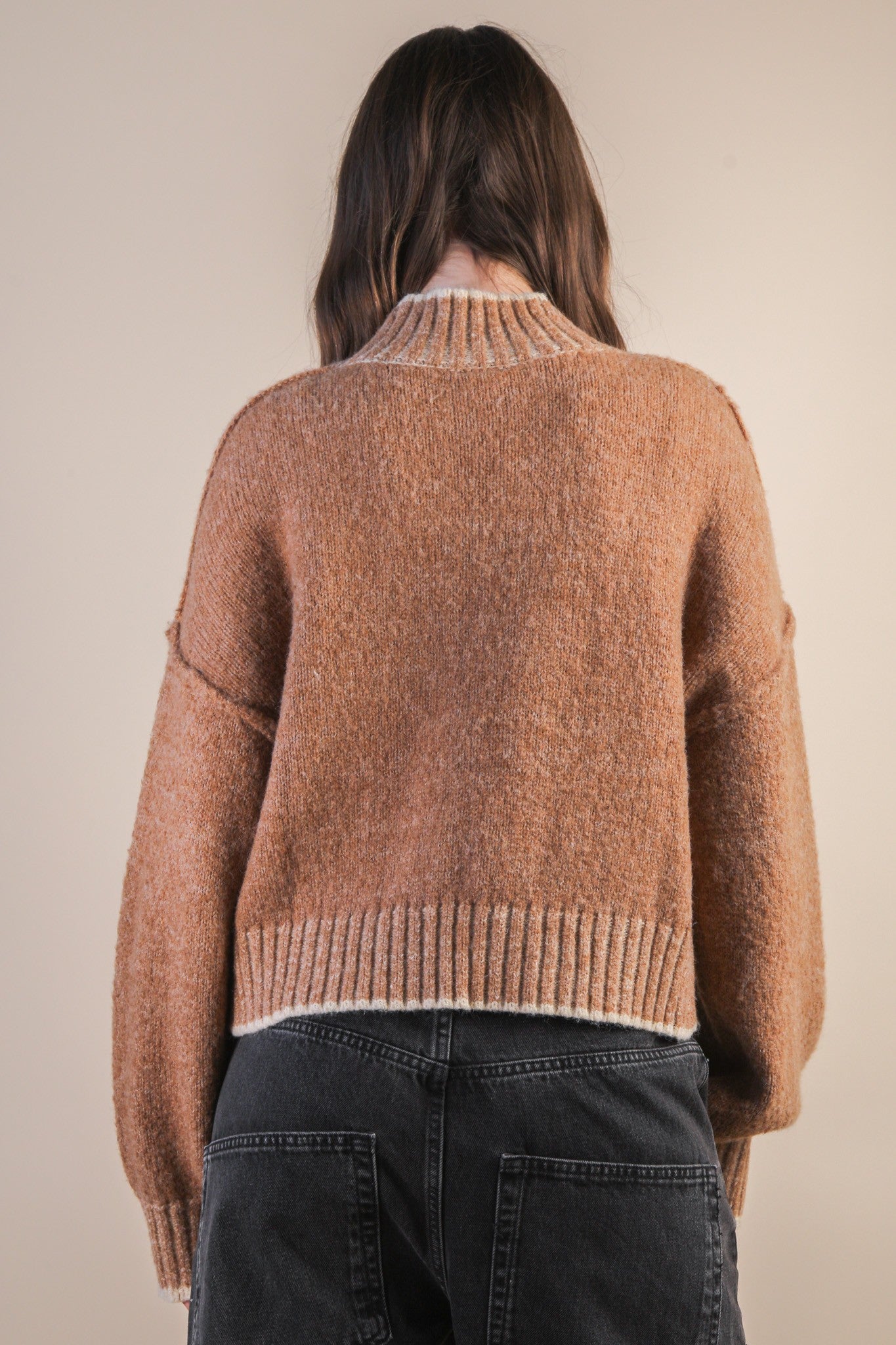 Back view of a woman wearing a mock neck mocha knit sweater with contrasting white trim against a plain background