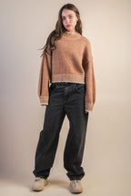 Woman wearing a mock neck mocha knit sweater with contrasting white trim and black jeans against a plain background