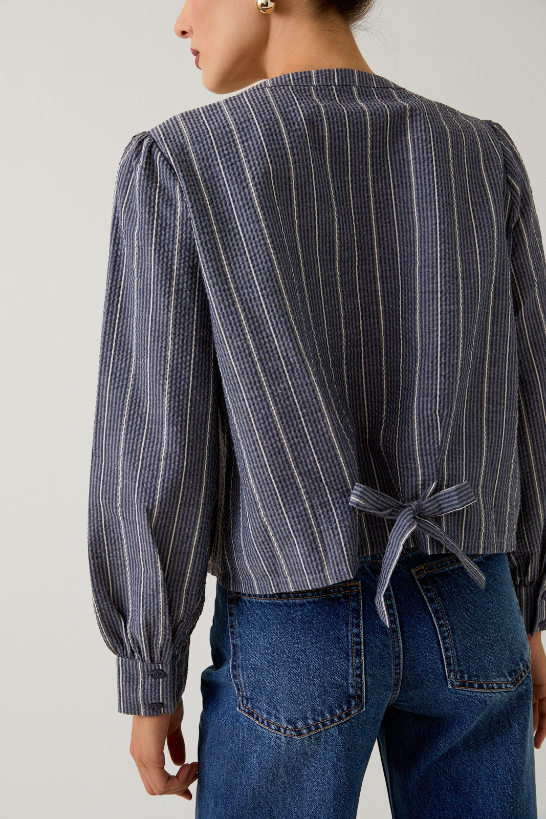 Back view of a woman wearing a navy striped blouse with tie back and jeans against a gray background