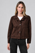 A woman modeling a dark brown suede jacket with a collar, button front, and two front patch pockets, paired with a white top and dark jeans, against a solid beige background.