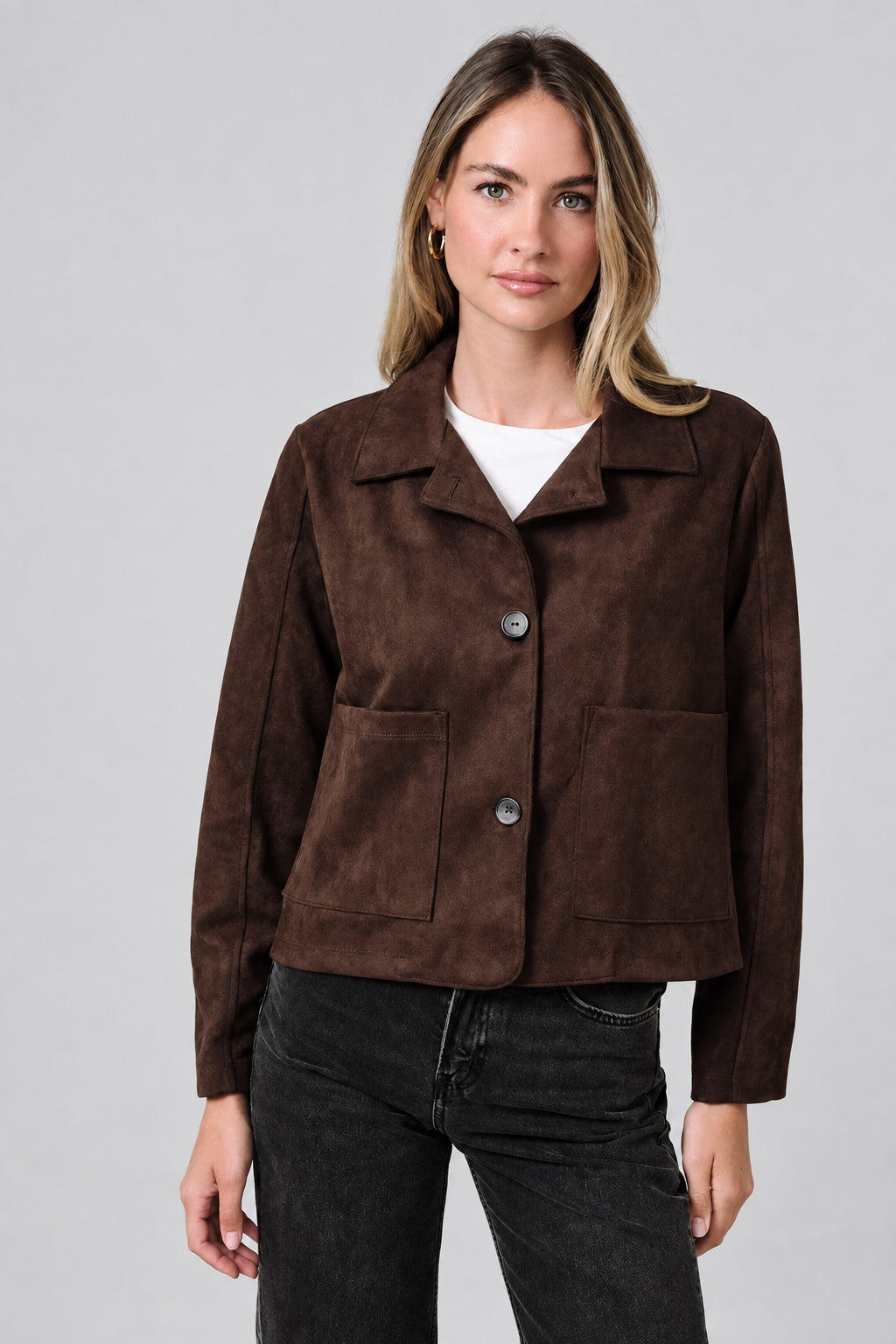 A woman modeling a dark brown suede jacket with a collar, button front, and two front patch pockets, paired with a white top and dark jeans, against a solid beige background.