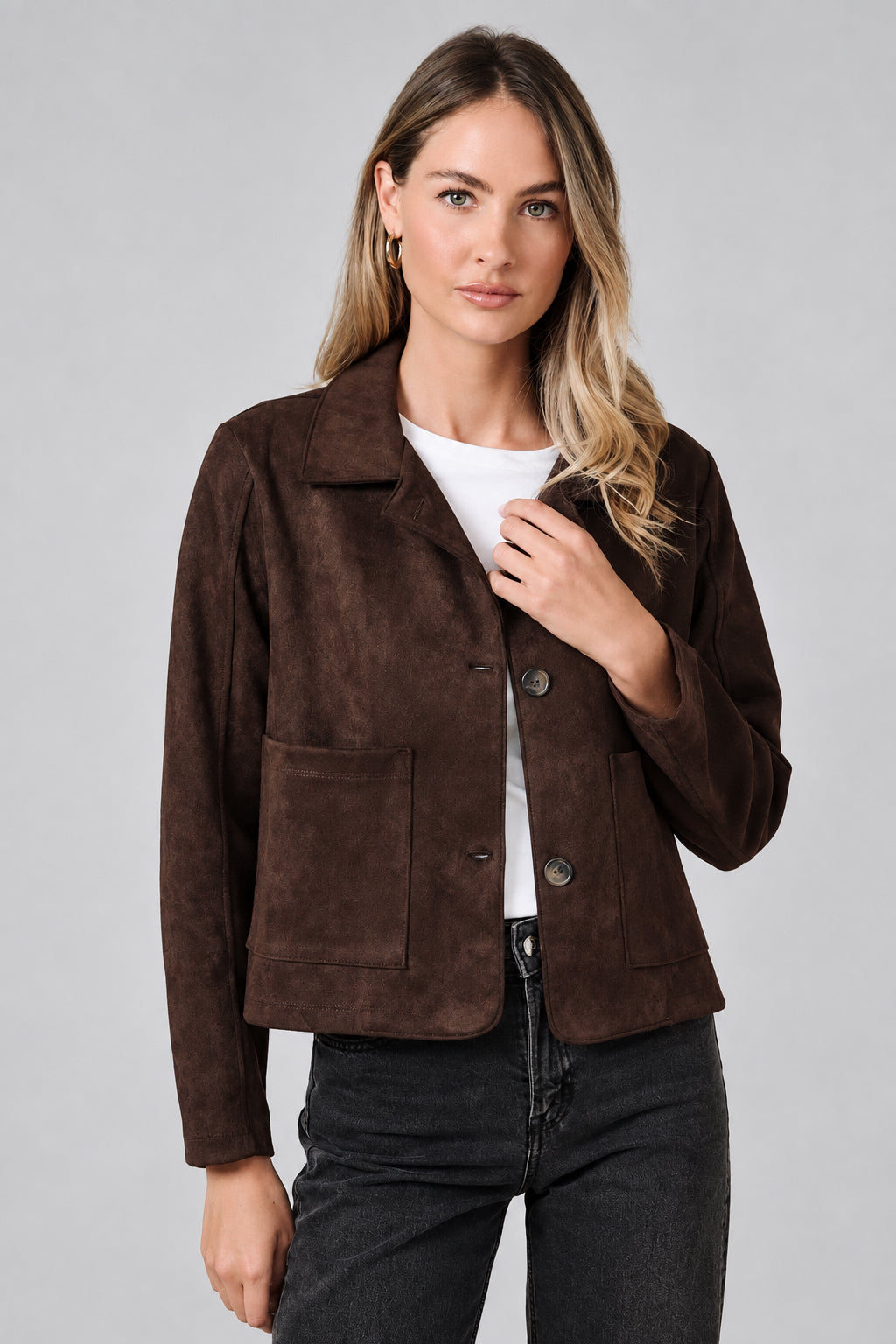 A woman modeling a dark brown suede jacket with a collar, button front, and two front patch pockets, paired with a white top and dark jeans, against a solid beige background.