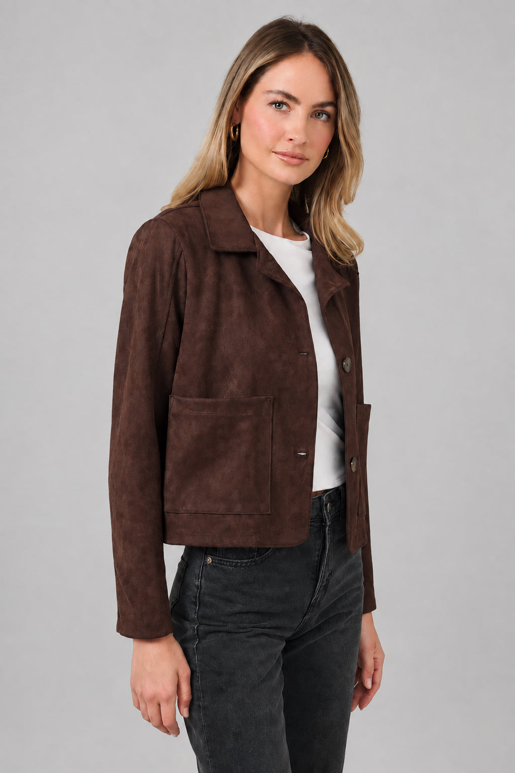 A woman modeling a dark brown suede jacket with a collar, button front, and two front patch pockets, paired with a white top and dark jeans, against a solid beige background.