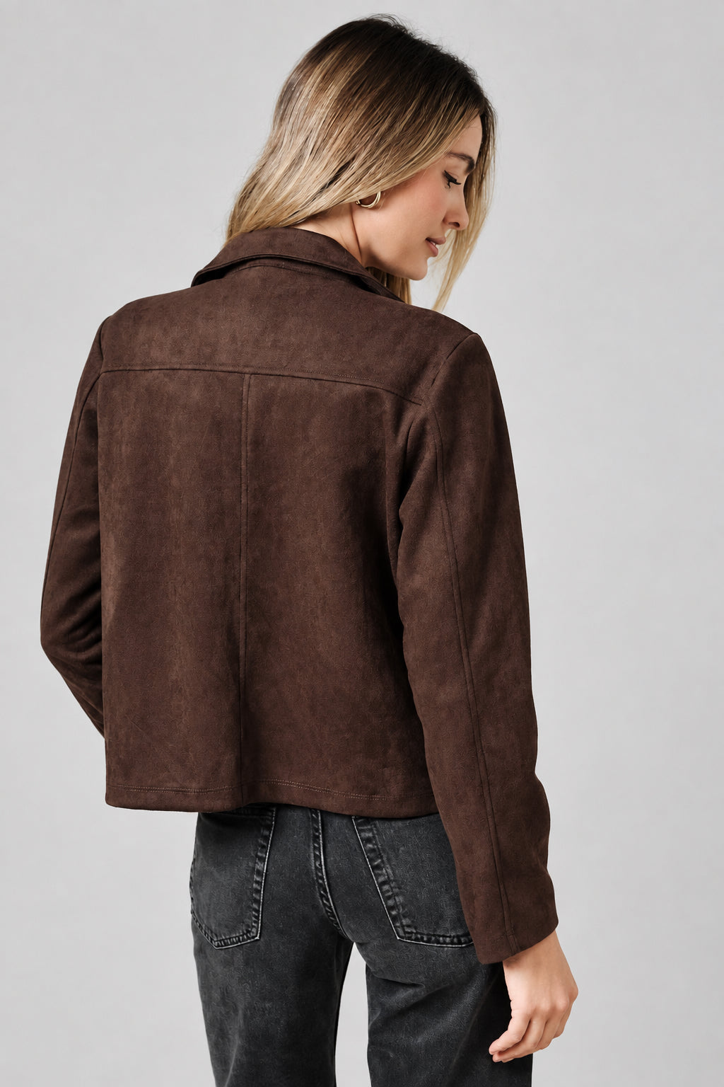 Back view of a woman modeling a dark brown suede jacket with a collar, button front, and two front patch pockets, paired with a white top and dark jeans, against a solid beige background.
