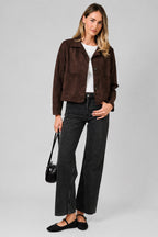 A woman modeling a dark brown suede jacket with a collar, button front, and two front patch pockets, paired with a white top and dark jeans, against a solid beige background.