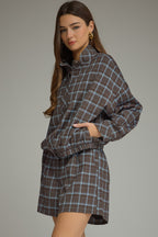 A woman modeling a brown and blue plaid long-sleeve jacket with matching shorts against a solid gray studio background.