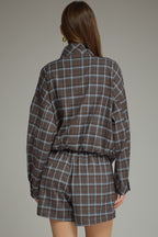 A woman shown from the back wearing a matching plaid long-sleeve jacket and shorts set in brown and blue  against a solid gray studio background.