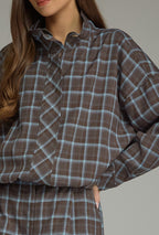 A woman modeling a matching plaid button-up jacket and shorts set in brown and blue. She is standing against a solid gray studio background.