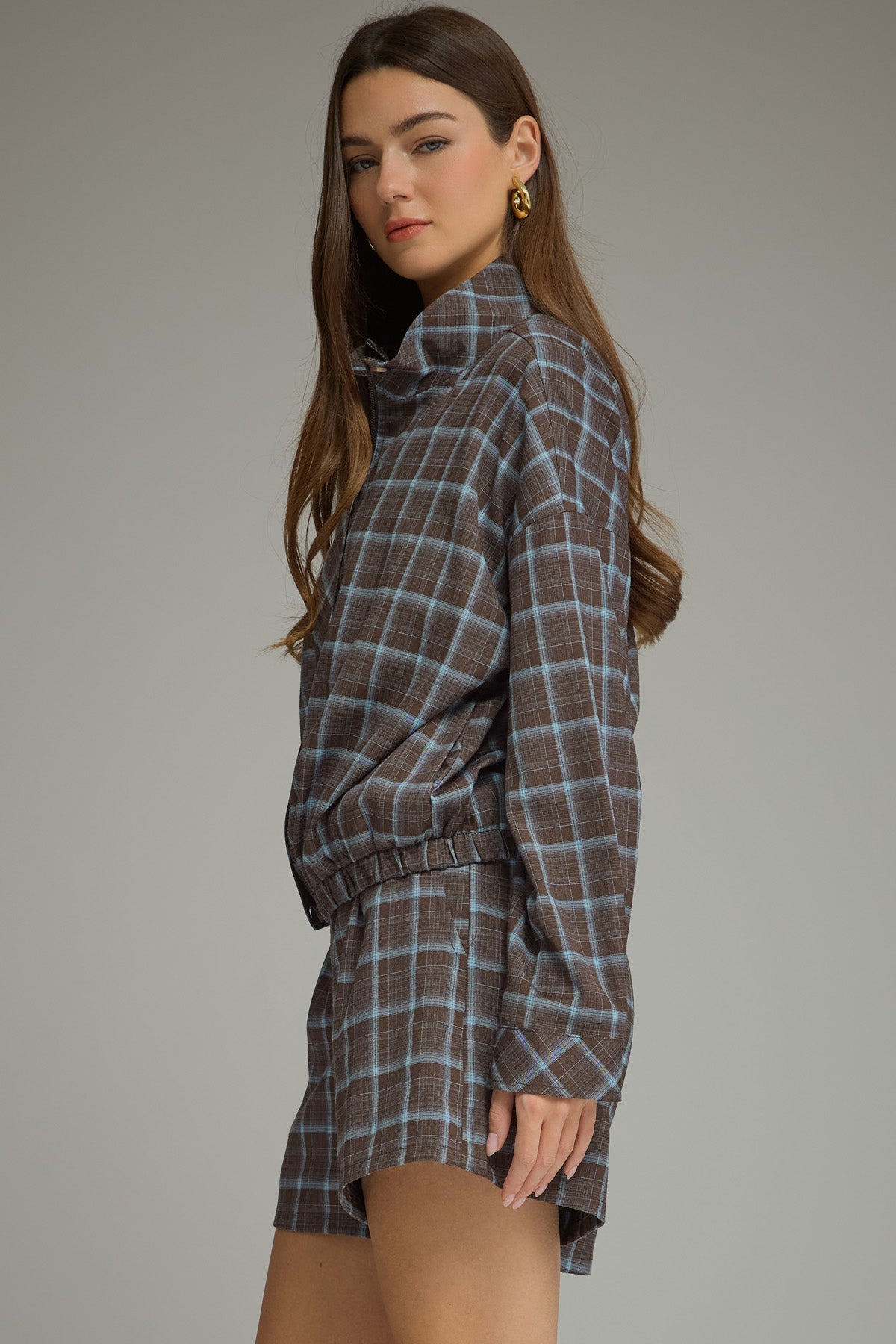 A woman modeling a matching plaid long-sleeve jacket and shorts set in brown and blue, photographed against a neutral gray studio background.