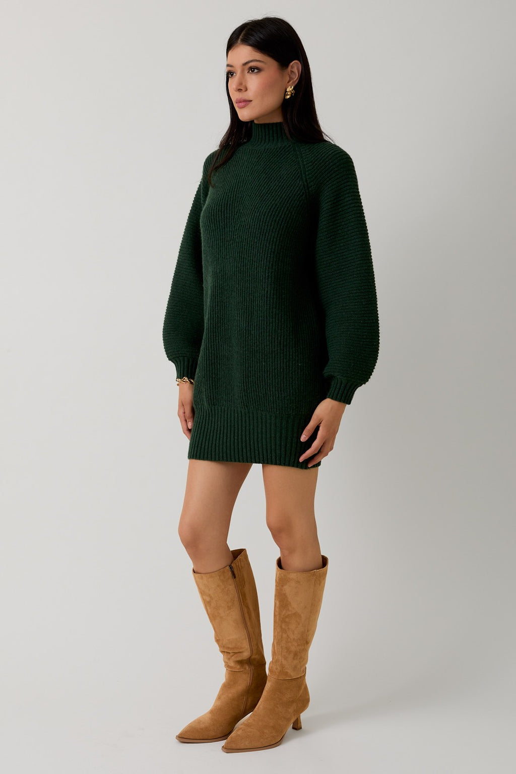 Woman wearing a hunter green sweater dress with camel boots against a plain background