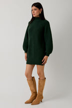 Woman wearing a hunter green sweater dress with camel boots against a plain background