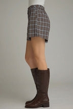 A side-profile view of a person wearing brown and blue plaid shorts with tan knee-high boots against a neutral gray studio background.