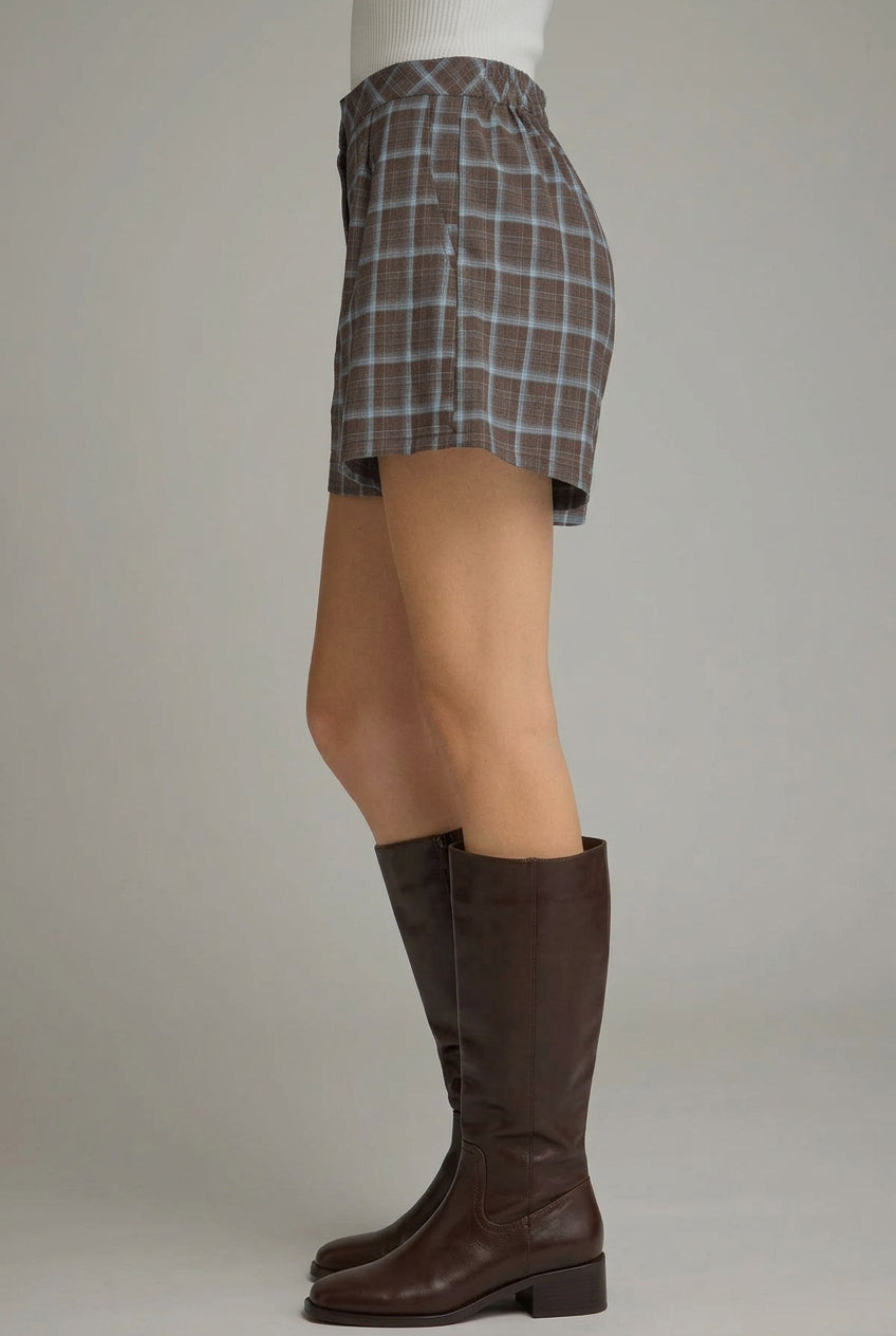 A side-profile view of a person wearing brown and blue plaid shorts with tan knee-high boots against a neutral gray studio background.