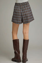 A person shown from the back wearing brown plaid shorts with a blue grid pattern and tan knee-high boots against a neutral gray studio background.