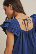 Woman wearing a midnight blue top with ruffled sleeves and tie back against a beige background