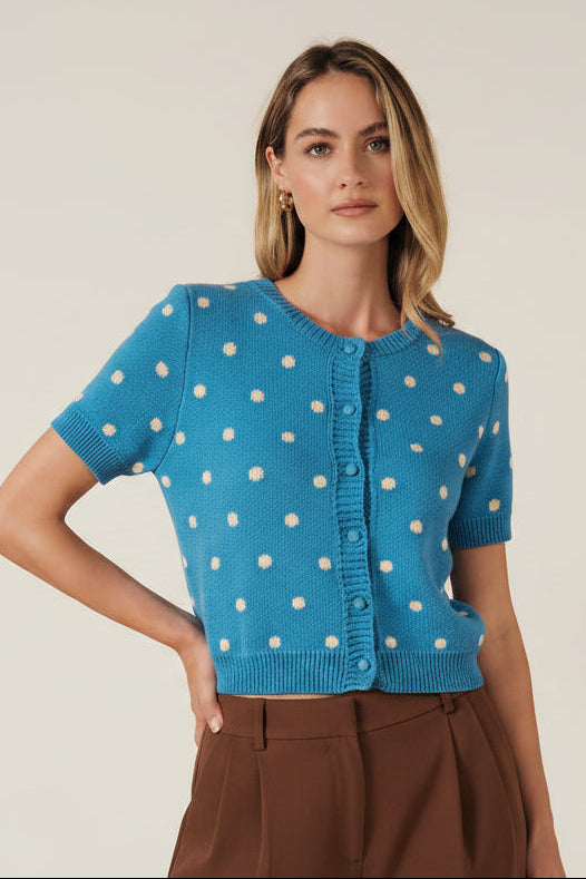 A woman modeling a bright blue short-sleeve knit cardigan with cream polka dots and a button-front closure, against a solid beige studio background.