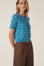 A woman modeling a bright blue short-sleeve knit cardigan with cream polka dots and a button-front closure, against a solid beige studio background.