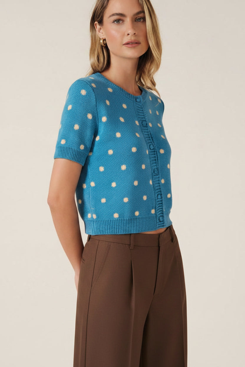 A woman modeling a bright blue short-sleeve knit cardigan with cream polka dots and a button-front closure, against a solid beige studio background.