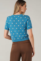 Back view of a woman modeling a bright blue short-sleeve knit cardigan with cream polka dots and a button-front closure, against a solid beige studio background.