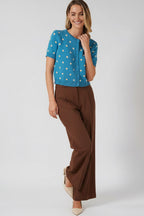 A woman modeling a bright blue short-sleeve knit cardigan with cream polka dots and a button-front closure, against a solid beige studio background.