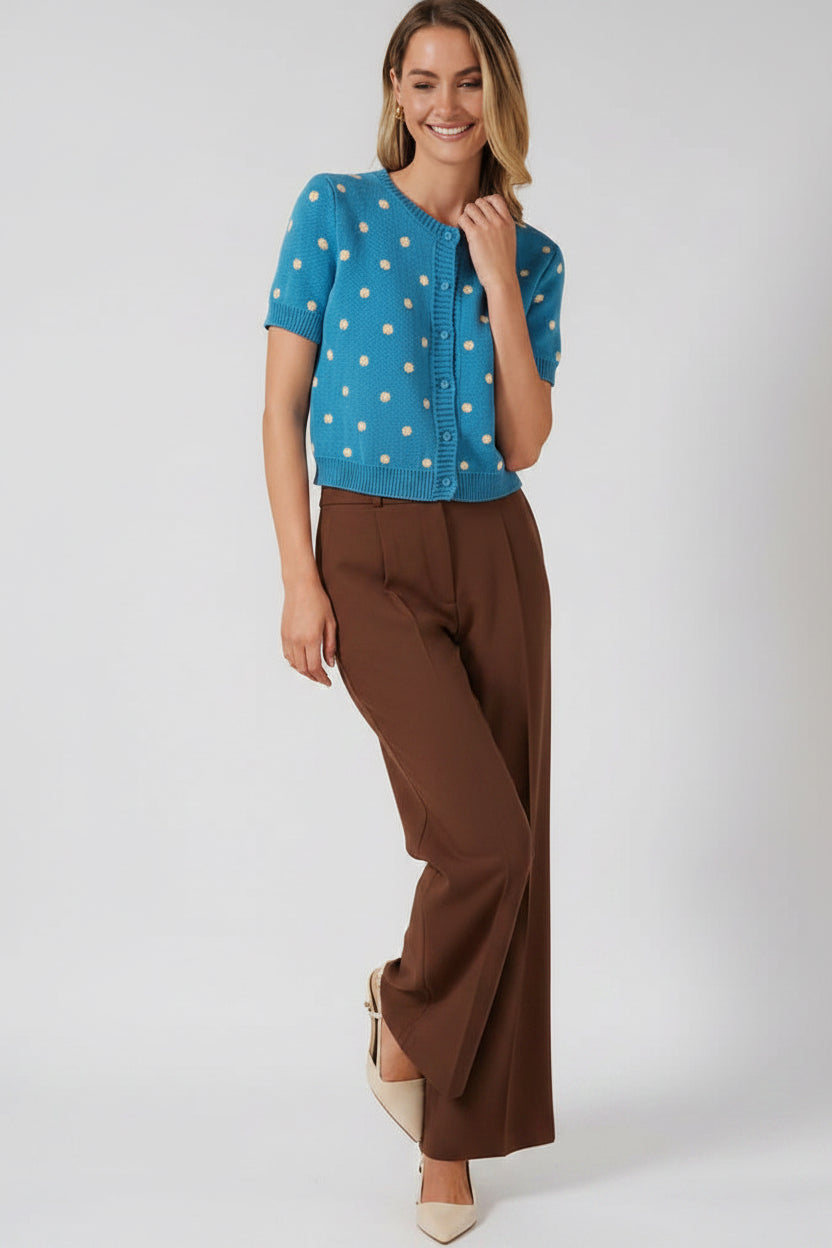 A woman modeling a bright blue short-sleeve knit cardigan with cream polka dots and a button-front closure, against a solid beige studio background.