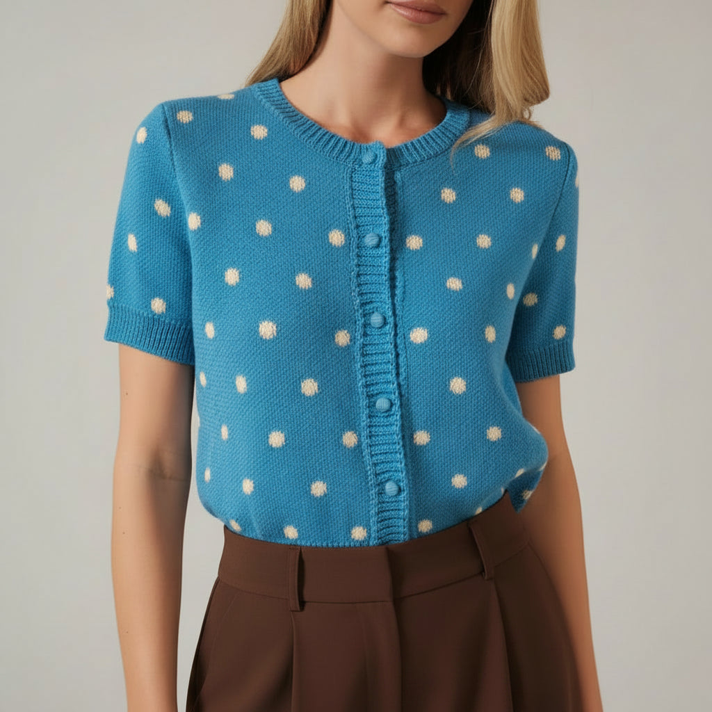 A woman modeling a bright blue short-sleeve knit cardigan with cream polka dots and a button-front closure, against a solid beige studio background.