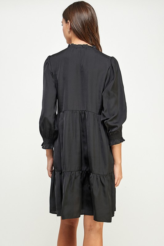Back view of a woman wearing a ruffle neckline tiered black satiny dress with long sleeves on a white background