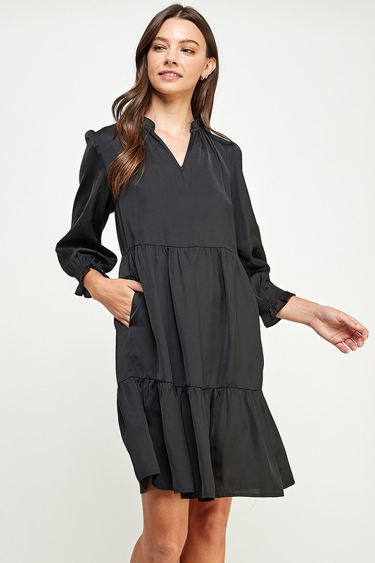 Woman wearing a ruffle neckline tiered black satiny dress with long sleeves on a white background