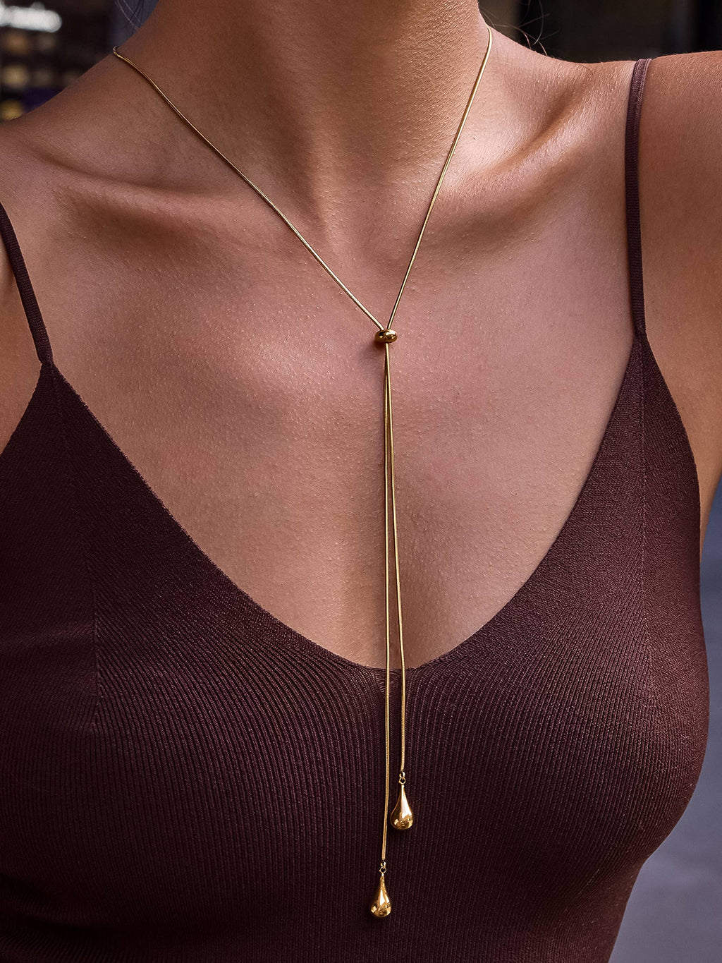 A close-up of a model wearing a delicate gold -plated lariat style drop teardrop pendant necklace styled over a dark brown ribbed camisole top.
