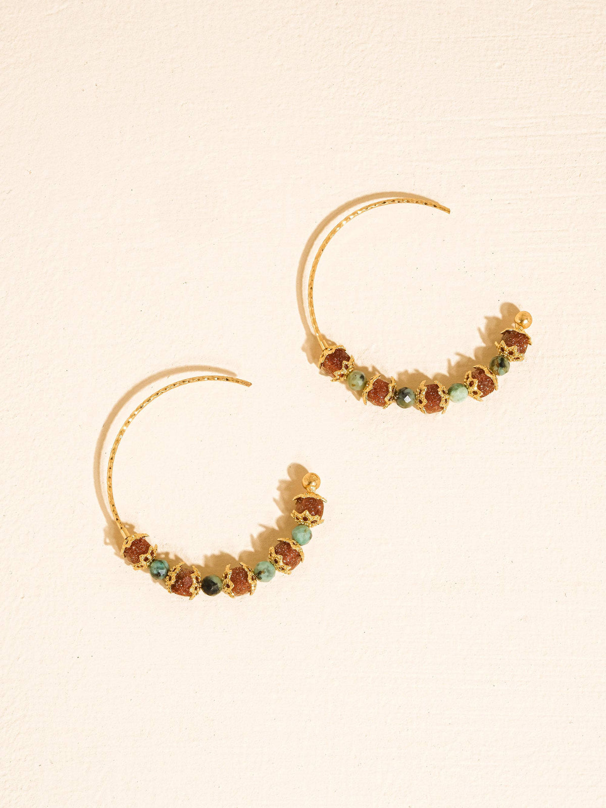 A pair of gold crescent-shaped hoop earrings decorated with small amber-brown and green faceted beads, displayed on a light beige textured background.