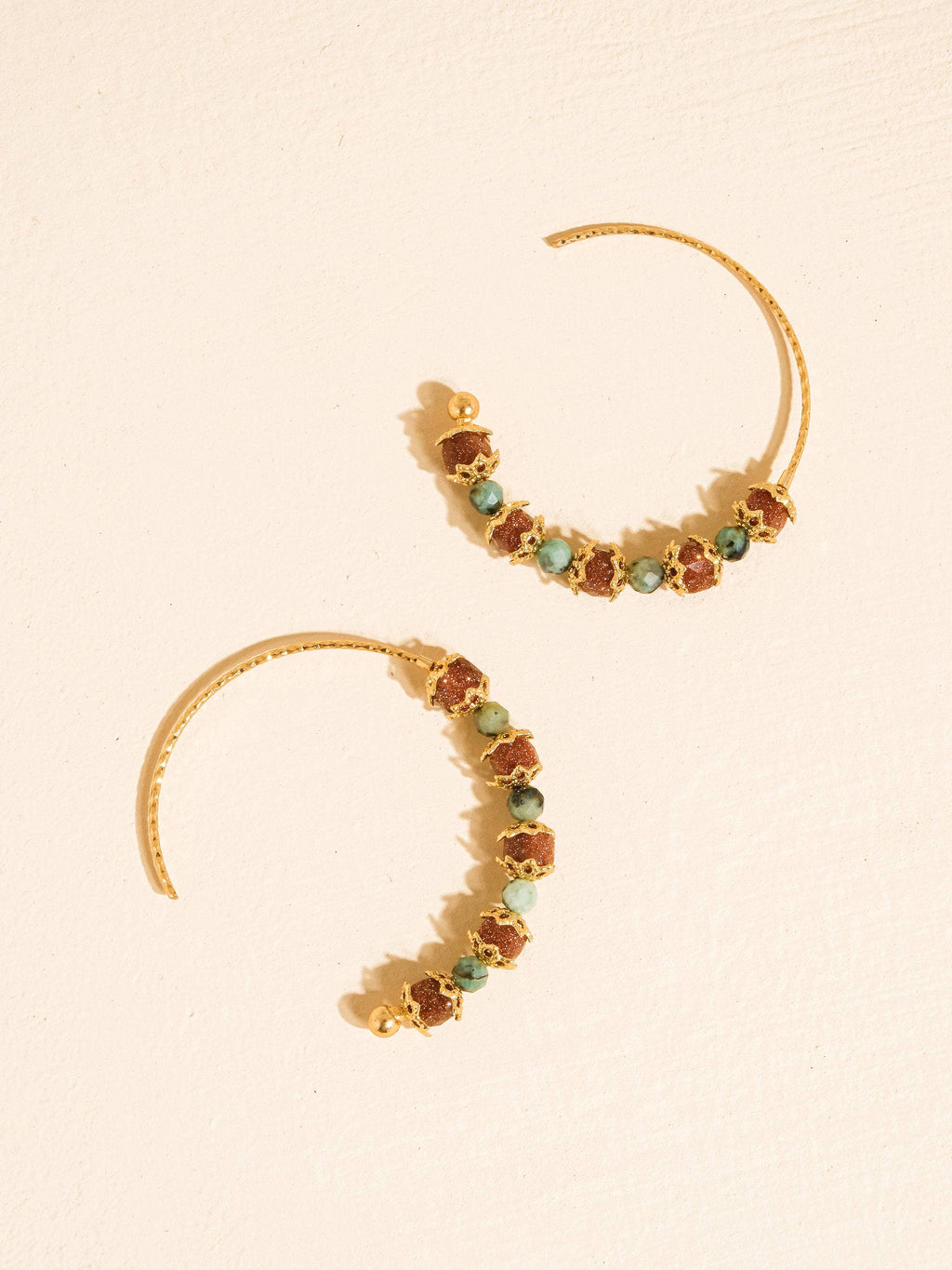 A pair of gold crescent-shaped hoop earrings decorated with small, irregular brown and green gemstones, displayed on a textured cream background.