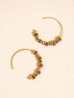 A pair of gold crescent-shaped hoop earrings decorated with small, irregular brown and green gemstones, displayed on a textured cream background.