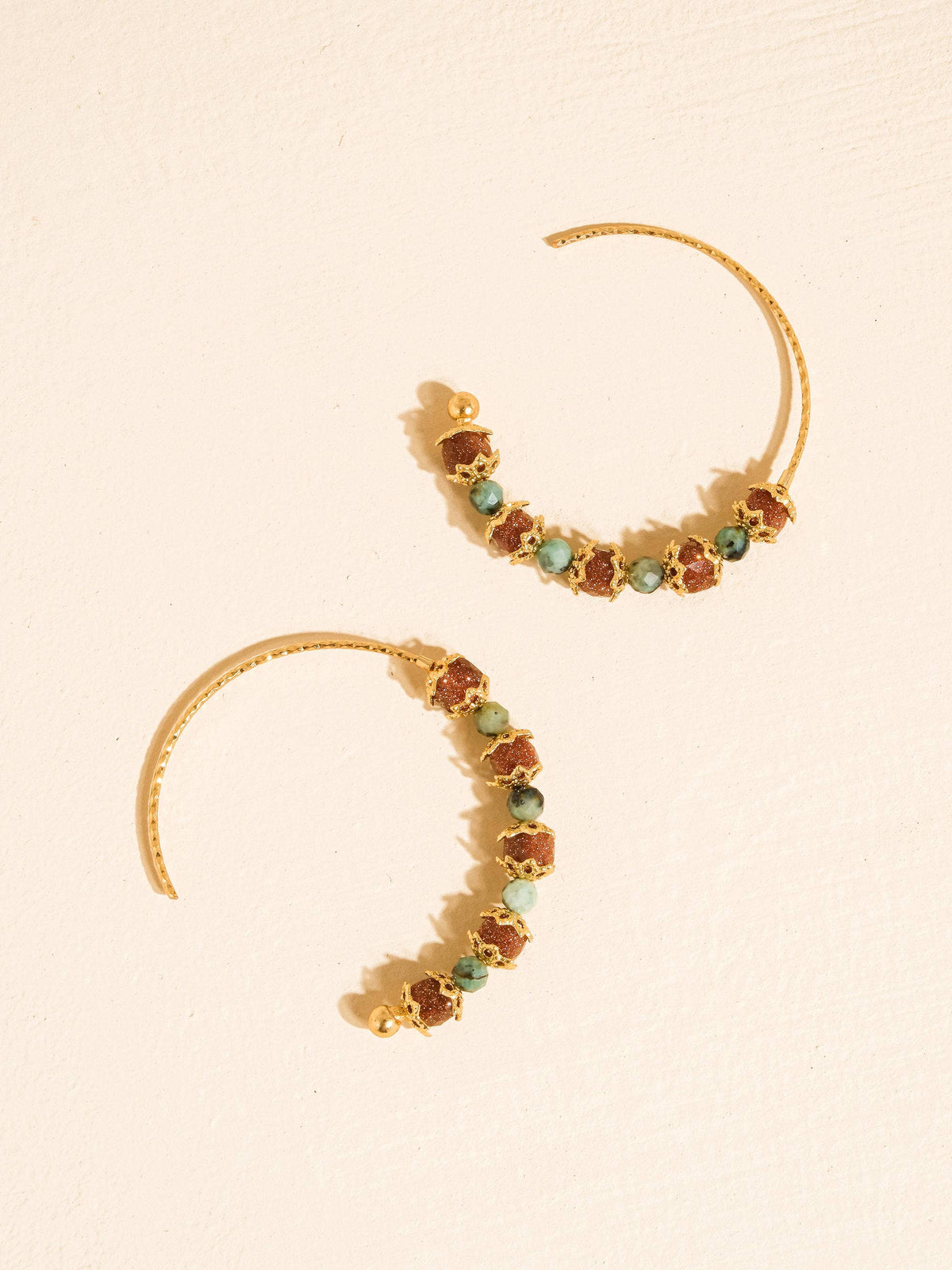 A pair of gold crescent-shaped hoop earrings decorated with small, irregular brown and green gemstones, displayed on a textured cream background.