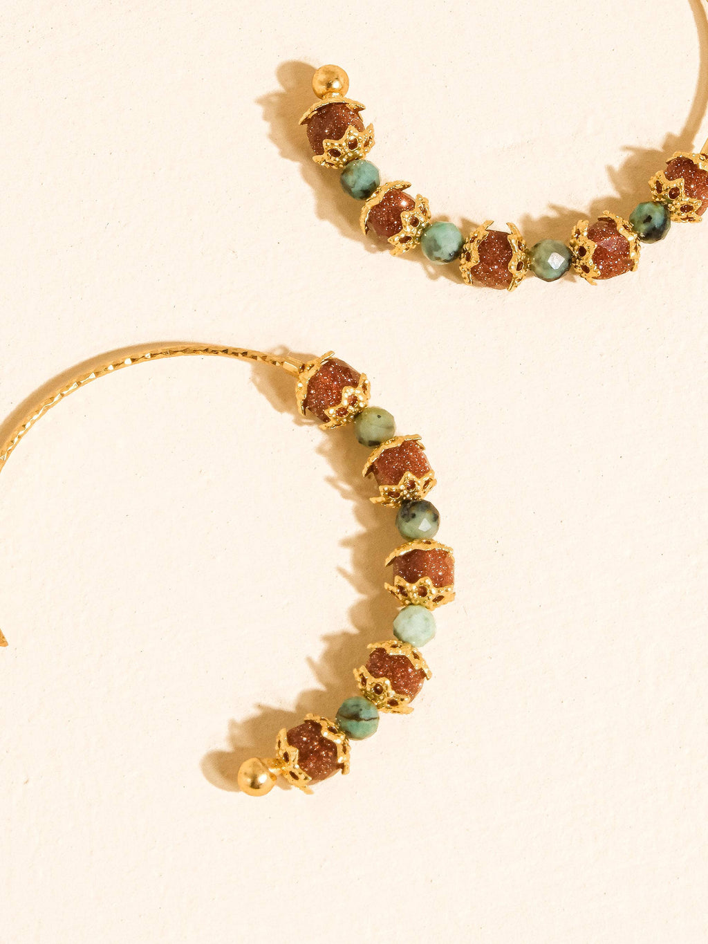 Gold hoop earrings with alternating textured brown and green faceted beads, displayed on a warm cream background.