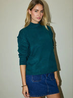 Woman wearing a hunter green sweater and blue denim skirt against a plain background