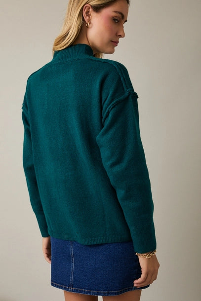 Back view of woman wearing a hunter green sweater and blue denim skirt against a plain background