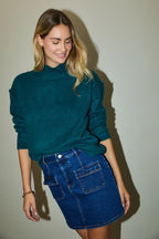 Woman wearing a hunter green sweater and blue denim skirt against a plain background