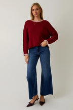 Woman wearing a berry sweater with cream contrast trim and jeans on a plain background